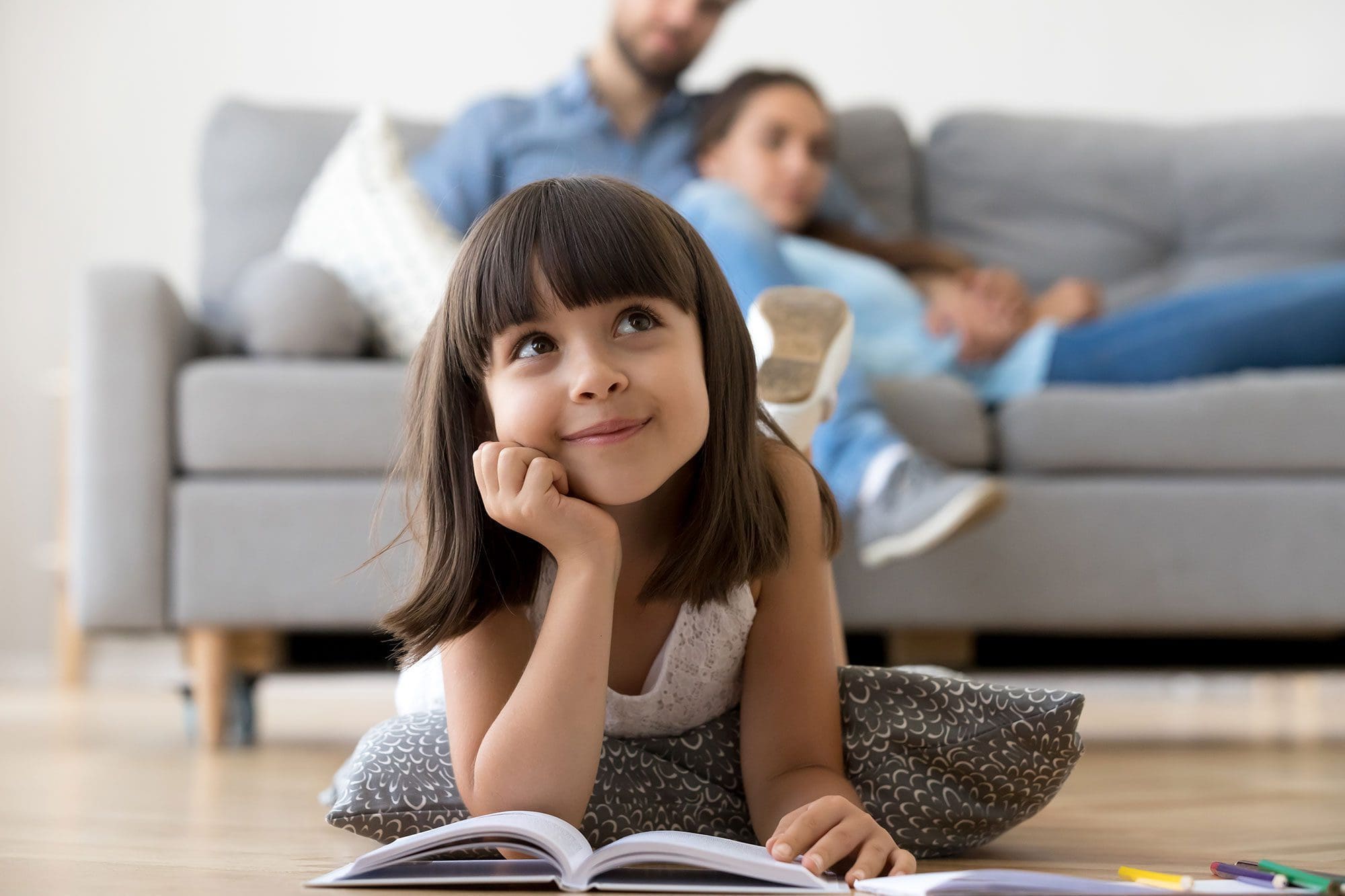 het beeld van een meisje dat op de grond ligt met een boek voor zich. Op de achtergrond liggen haar ouders op de bank.